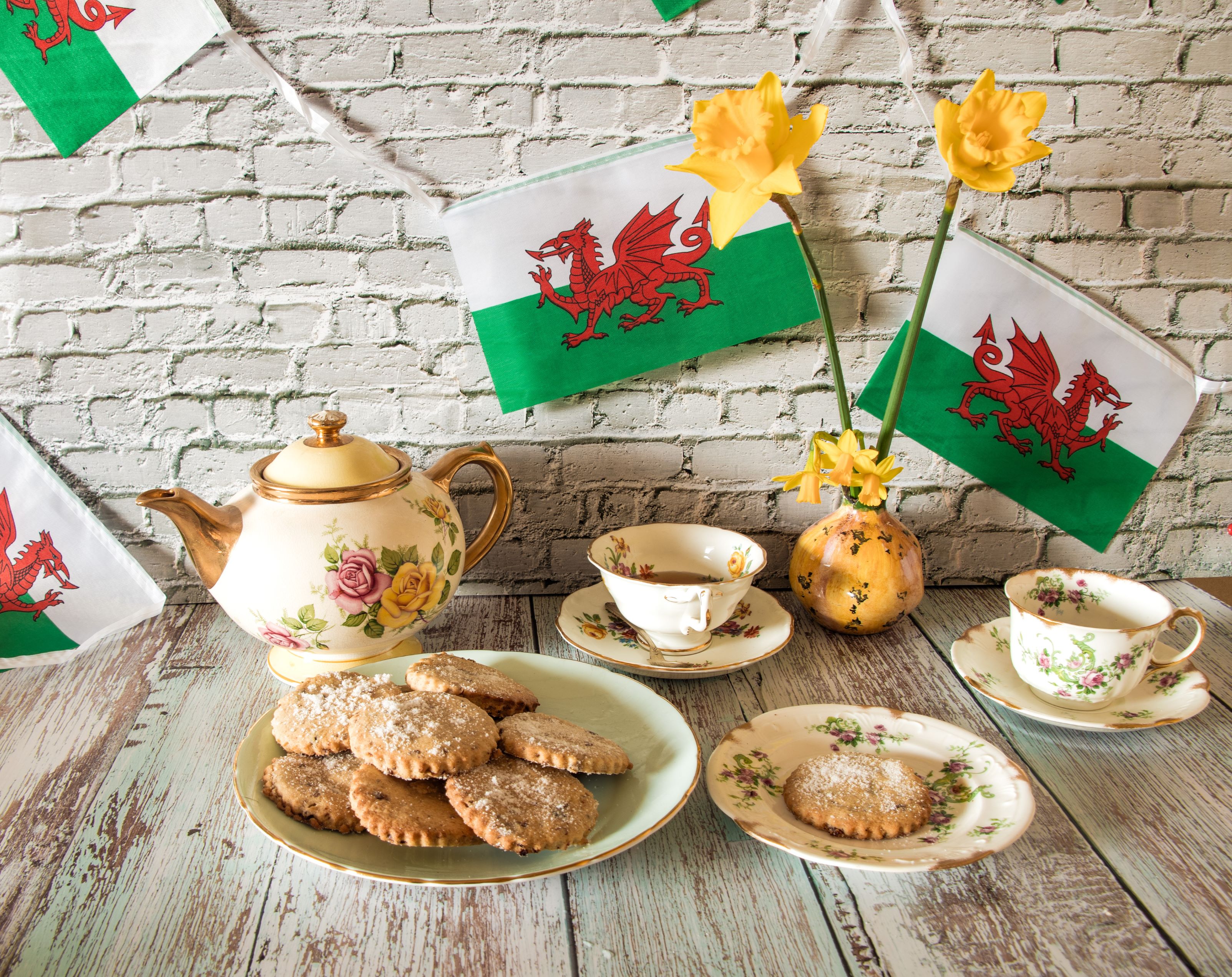 Welsh products, including Daffodils and Welsh cakes as part of a tea set with flag of Wales in background 