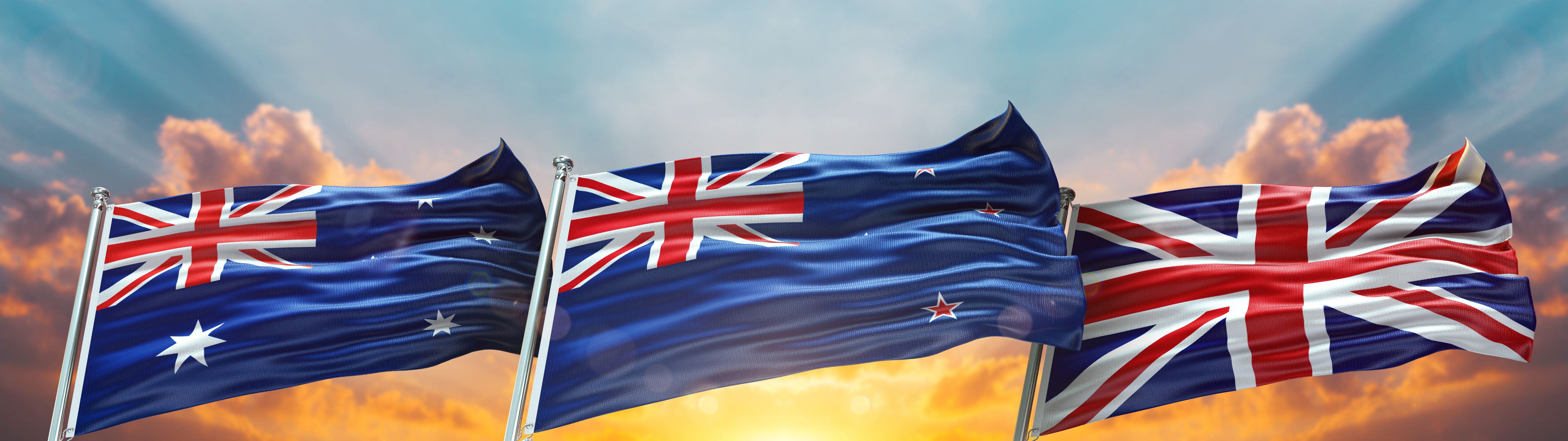 UK, New Zealand and Australia flags on flagpoles with sun lit sky behind